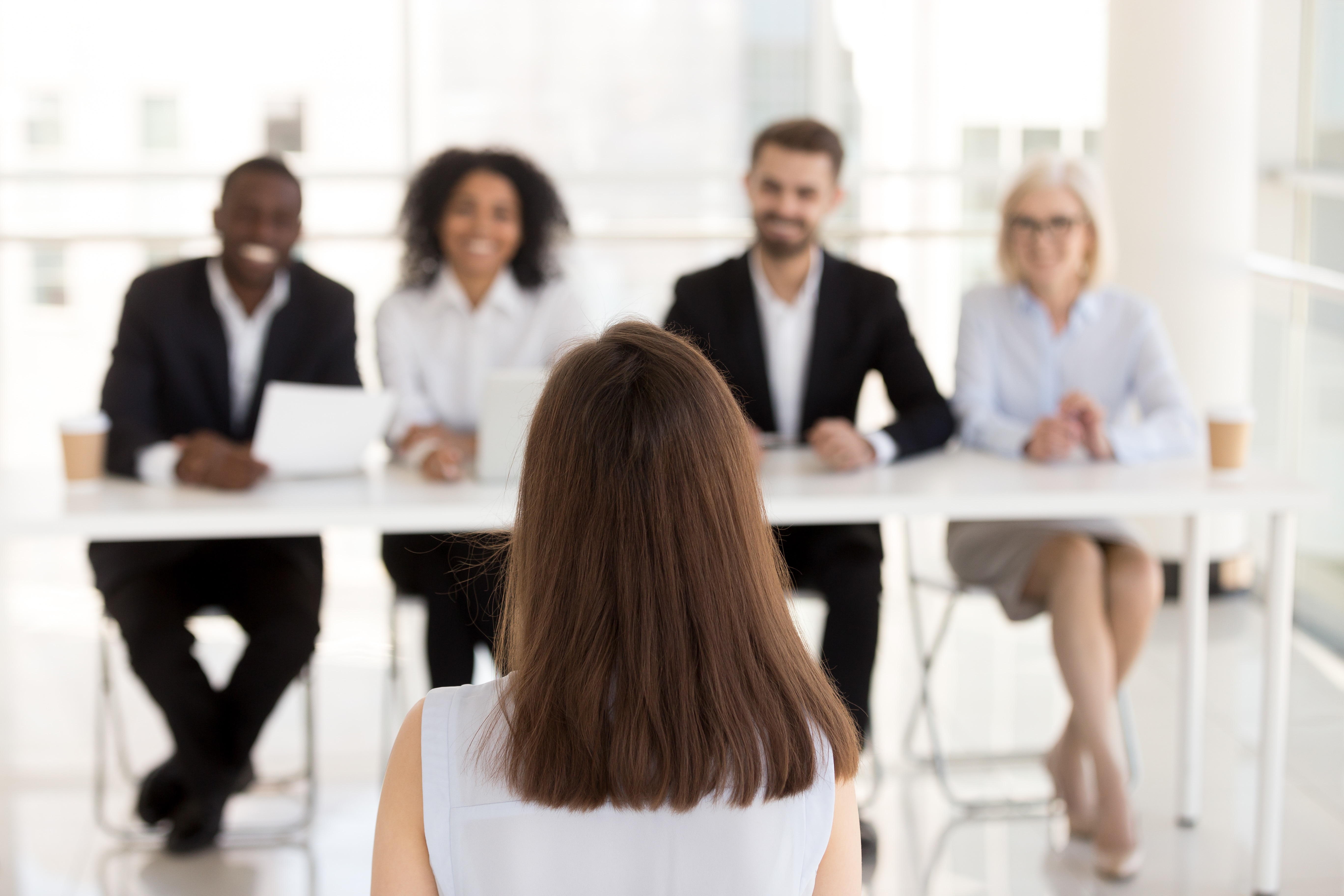  A woman sits with her back to the camera in front of a diverse hiring panel smiling and looking at her.