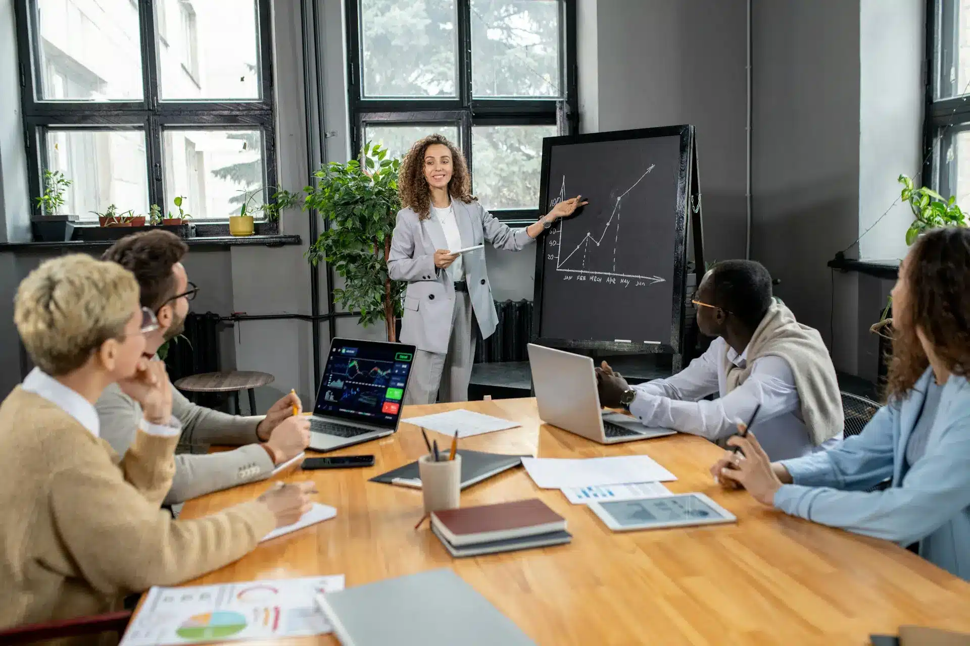 A diverse team of professionals in a modern meeting room, attentively listening to a female presenter standing by a blackboard with a graph showing upward trends. The presenter, dressed in a light grey suit, is explaining data while pointing at the chart. The group, seated around a wooden table, includes men and women of different ethnicities, with laptops and documents spread out, suggesting a collaborative and analytical business environment.